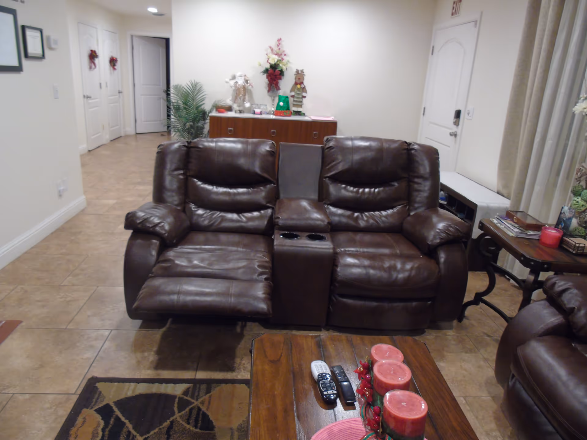 A cozy living room area with a brown leather reclining loveseat, a wooden coffee table with three red candles and remote controls, and a side table with magazines and a red candle. In the background, there is a wooden cabinet decorated with holiday figurines and a potted plant. The room has tiled flooring and light-colored walls.
