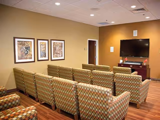 A small common area with multiple patterned armchairs arranged in rows facing a wall-mounted flat screen TV. The room has wood flooring, beige and brown walls, and three framed abstract artworks on one wall.