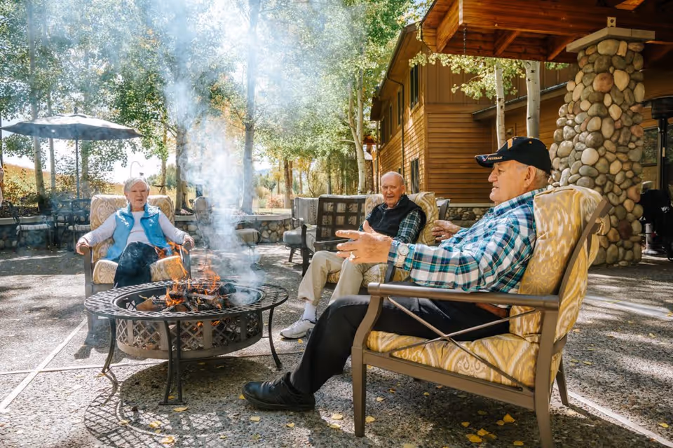 Three people sit around a smoking fire pit on a shaded outdoor patio beside a wood-sided lodge with trees and stone columns.