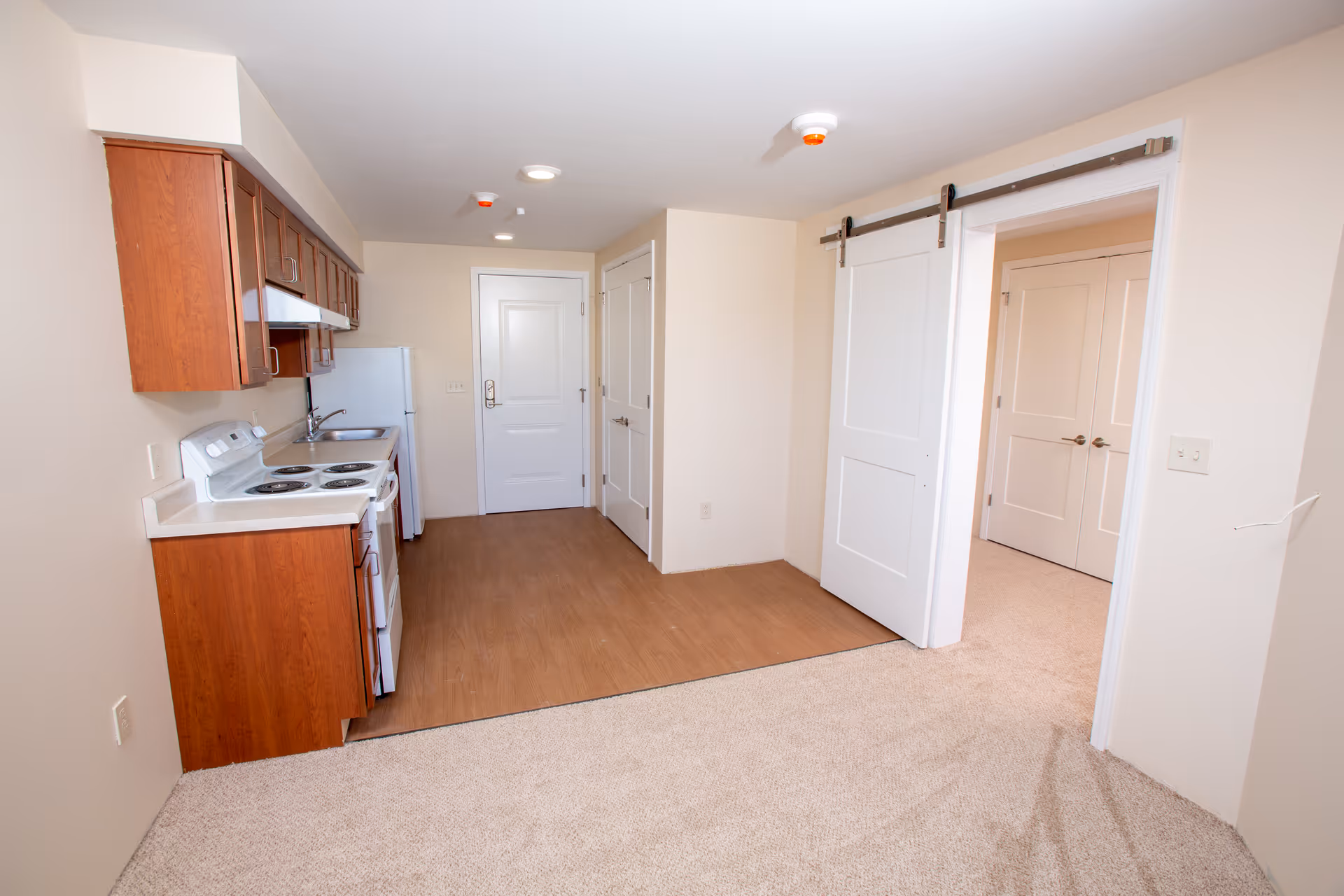 Interior view of a small kitchen area with wooden cabinets, a white electric stove, a sink, and a refrigerator. The kitchen floor is wood laminate, transitioning to carpet in the adjacent room. There is a white sliding barn door on a metal track leading to another room with double doors. The walls are painted light beige, and the ceiling has recessed lighting and smoke detectors.