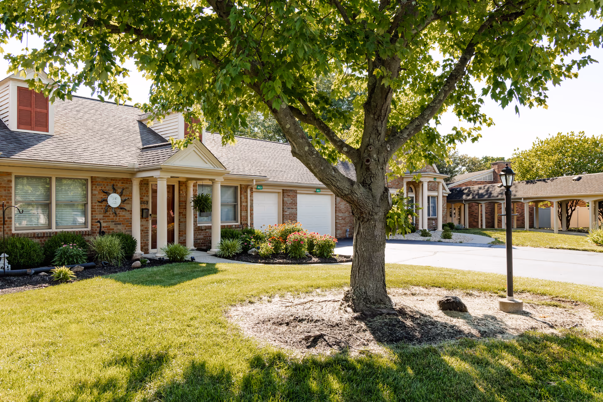 Exterior view of Westover Retirement Community showing a brick building with white columns at the entrance, a large tree in the foreground, well-maintained green lawn, shrubs, and a lamp post on a sunny day.