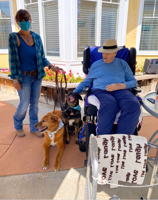 An elderly man wearing a hat and blue clothes sits in a wheelchair outside near a garden bed with flowers. A woman wearing a face mask, sunglasses, and casual clothes stands beside him holding leashes for two dogs, one brown and one black. The setting appears to be a sunny outdoor patio area of a senior living facility.
