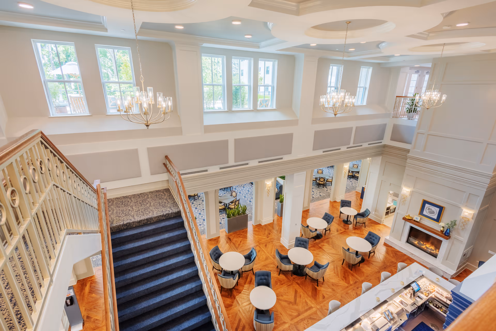 View from the top of a staircase looking down into a spacious, elegant common area with round tables and blue cushioned chairs arranged on a polished wooden floor. The room features large windows, chandeliers, and a fireplace with decorative items on the mantel. The area appears bright and welcoming with natural light coming through the windows.