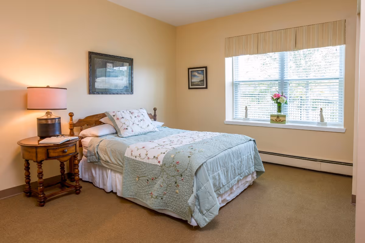 A cozy bedroom with a neatly made bed featuring a light blue and floral quilt. There is a wooden nightstand with a lamp and a book on it next to the bed. The room has beige walls, a large window with blinds, and a vase with flowers on the windowsill. Two framed pictures hang on the walls.