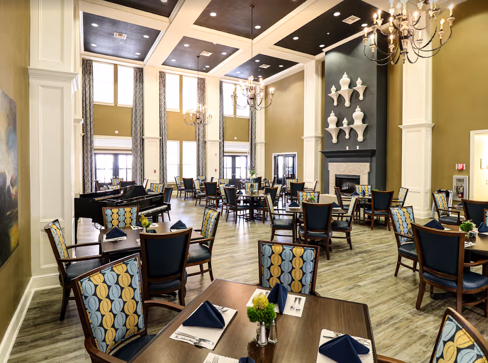Large elegant dining room with multiple set tables and patterned chairs, chandeliers, tall windows, a fireplace and a grand piano.