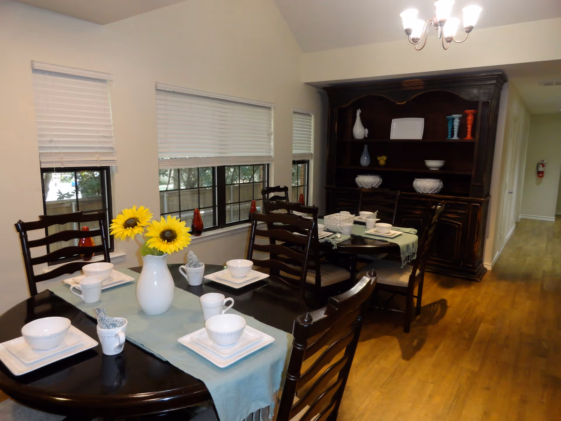 Dining room with two dark wooden tables set with white dishes, cups, and napkins. A white vase with two sunflowers is placed on the table in the foreground. The room has three windows with white blinds and a large dark wooden cabinet with decorative items on shelves. Hardwood flooring and a chandelier light fixture are visible.