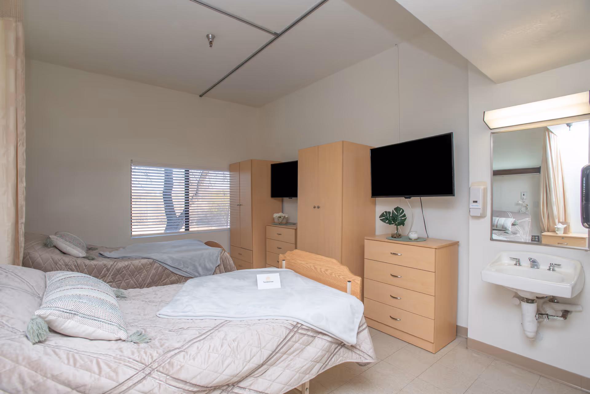Two single beds in a senior living bedroom with light-wood dressers, a wall-mounted TV, a window with blinds, and a sink with a mirror.
