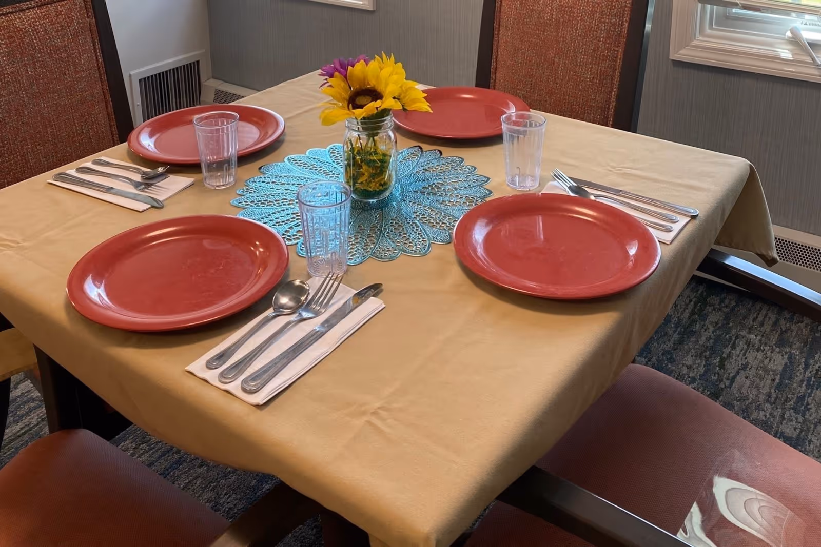 A dining table set for four with red plates, clear drinking glasses, and silverware on white napkins. The table is covered with a beige tablecloth and has a blue lace doily centerpiece with a small jar holding sunflowers and other flowers. The room has carpeted flooring and a window with blinds.