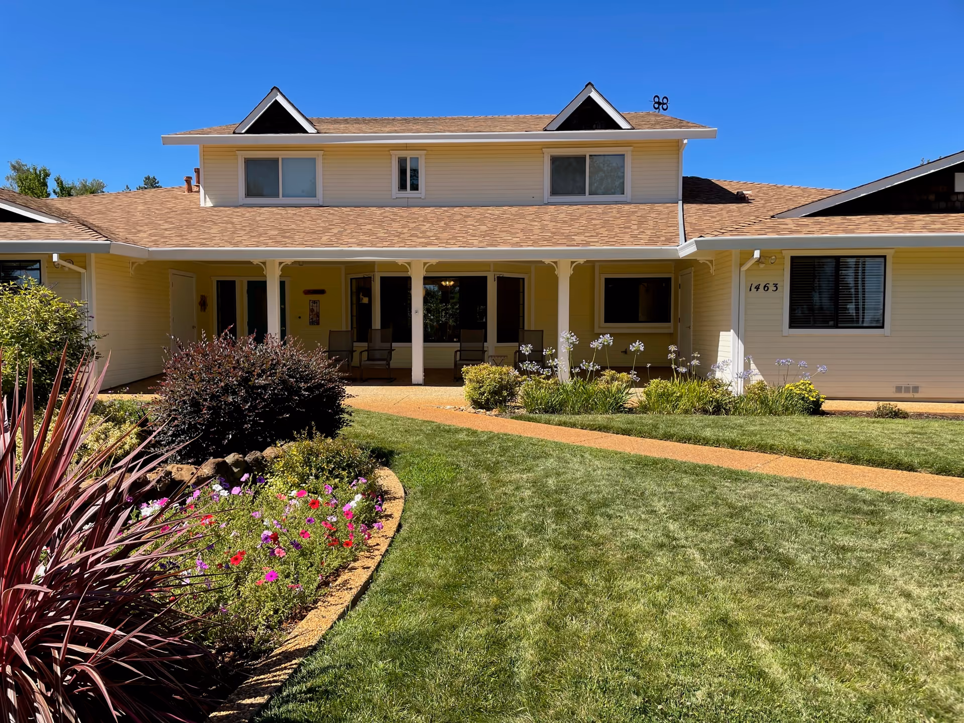 Front exterior of a two-story yellow house with a covered porch, walkway, lawn, and flowerbeds.