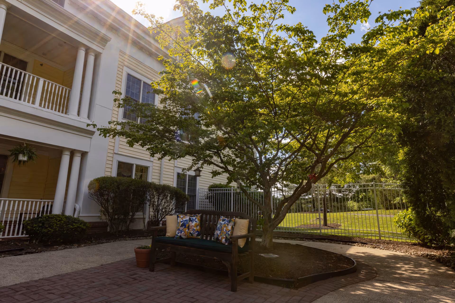 Outdoor courtyard area at Mira Vie at East Brunswick featuring a wooden bench with colorful cushions under a leafy tree, surrounded by a paved walkway, bushes, and a metal fence with a building in the background.