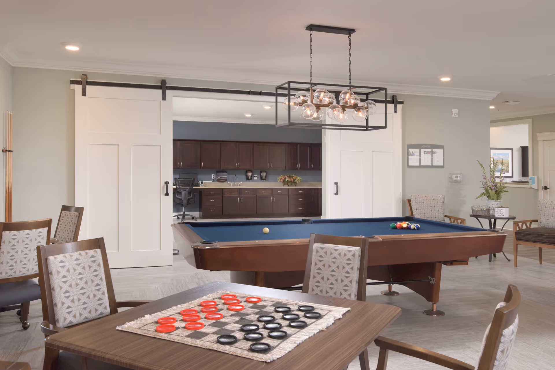 A bright and spacious game room featuring a pool table with balls arranged for a game, a table with a checkers board set up with red and black pieces, several cushioned chairs, and a modern light fixture hanging from the ceiling. In the background, there are sliding white barn doors partially open revealing a room with dark wooden cabinets and a desk chair.