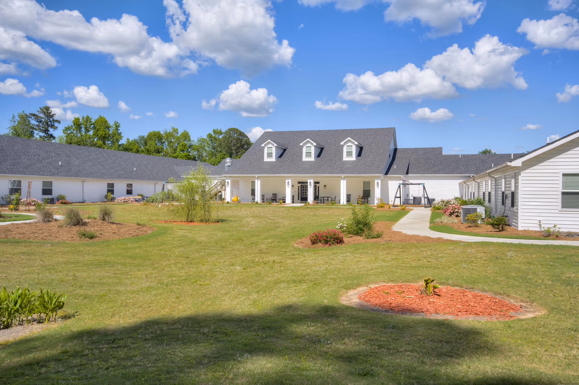 A large, single-story white building with a dark gray roof surrounded by a well-maintained lawn and landscaped garden beds under a blue sky with scattered clouds.