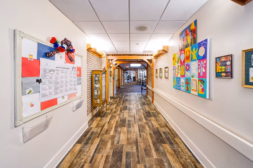 A well-lit hallway in a senior living facility with wood-patterned flooring and white walls. The left wall features a bulletin board with a calendar and decorations, while the right wall displays colorful artwork and framed pictures. Wooden archways and a glass display cabinet are visible along the corridor, with chairs and additional decorations further down the hall.