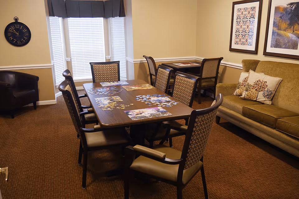 A cozy room with a rectangular wooden table surrounded by eight cushioned chairs. Several puzzle pieces and puzzle boards are spread out on the table. The room has beige walls with white trim, a green sofa with decorative pillows featuring tree designs, two framed pictures on the wall, a black armchair in the corner, and a large window with blinds and a dark valance. A round black wall clock is mounted on the wall near the window.