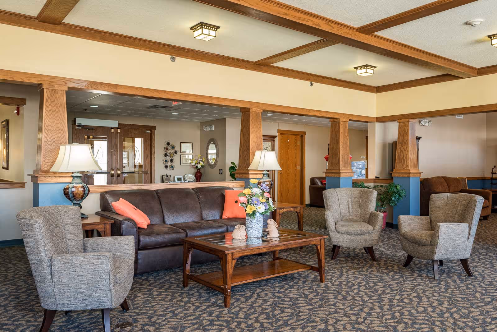 Community lounge with a leather sofa, four upholstered armchairs, a wooden coffee table, lamps and floral arrangements under coffered ceilings.