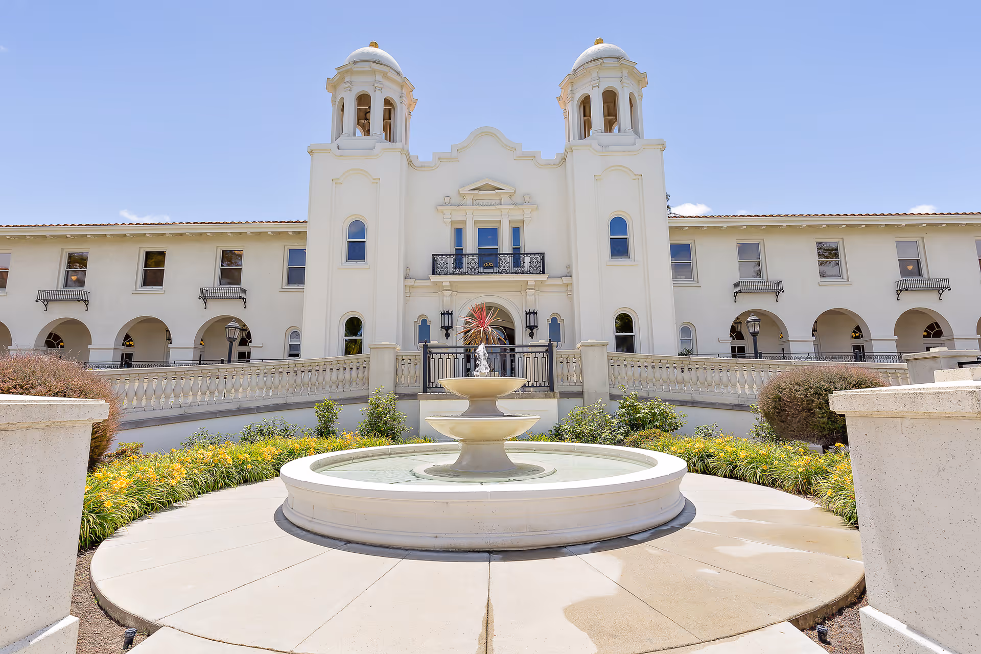 Mediterranean-style white retirement community building with twin towers and a circular fountain in the landscaped front courtyard.