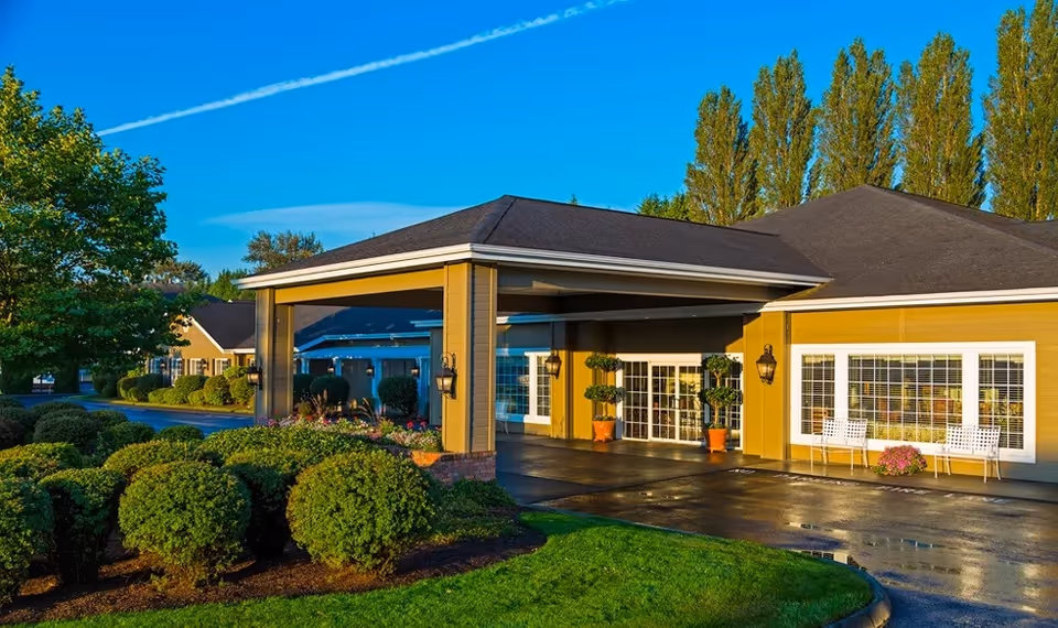 Exterior view of Canterbury Gardens Memory Care Community building with a covered entrance, manicured bushes, green lawn, and tall trees under a clear blue sky.