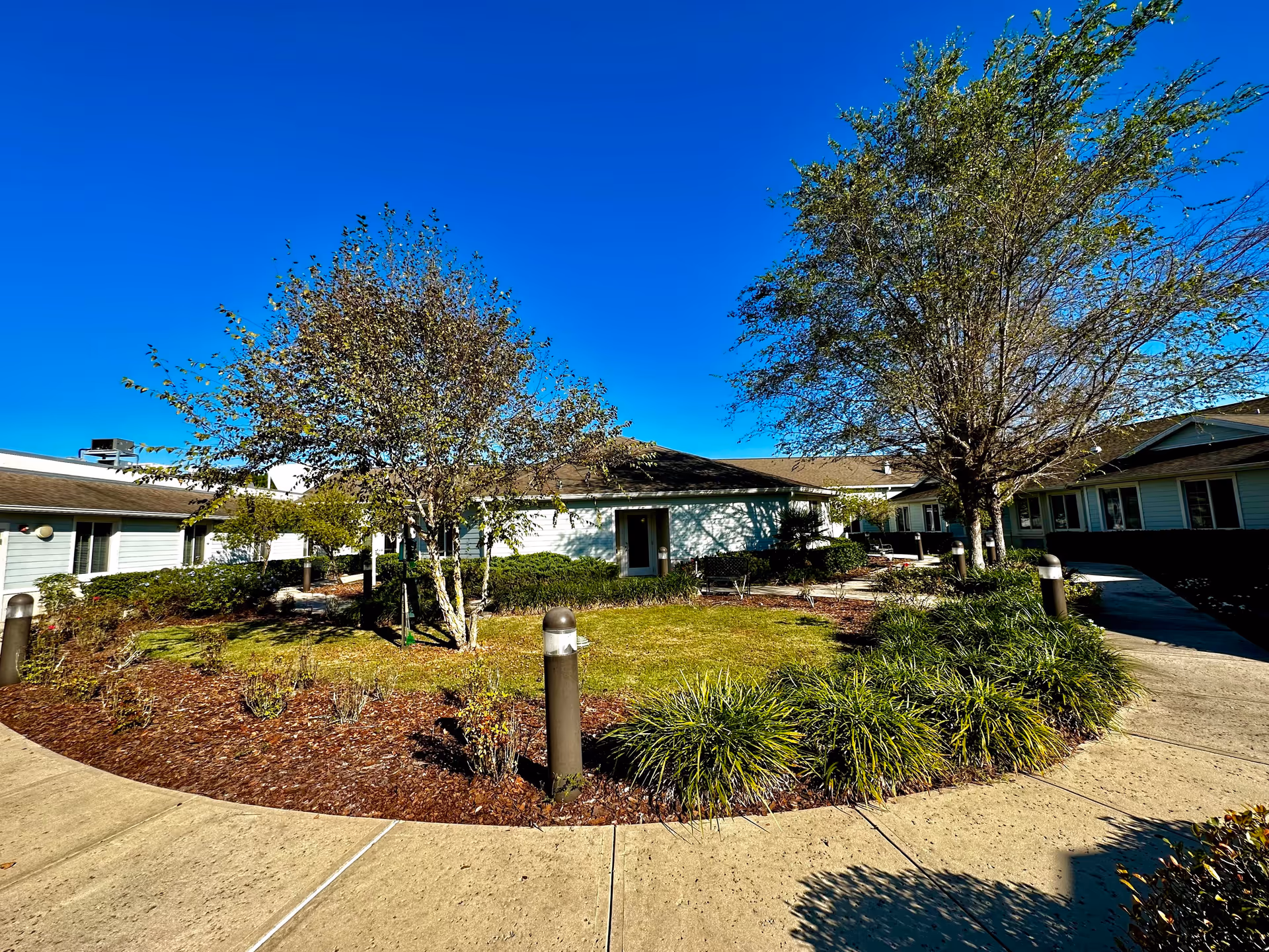 Outdoor courtyard area of a senior living facility with a circular walkway surrounding a landscaped garden featuring trees, bushes, and grass under a clear blue sky. The single-story building with light-colored siding and multiple windows surrounds the courtyard.