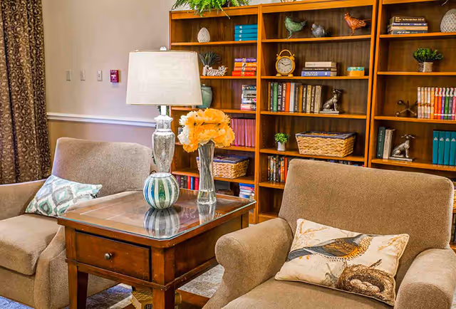 Two upholstered armchairs flank a wooden side table with a lamp and vase of flowers in front of a large bookshelf filled with books and decor.