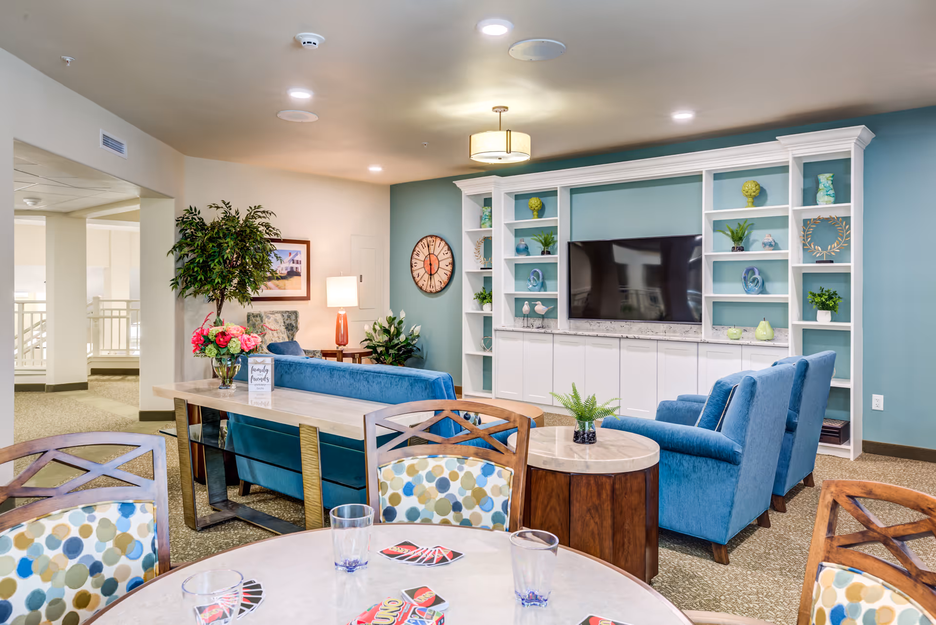 A cozy living room area in a senior living facility with blue upholstered sofas and armchairs arranged around a large flat-screen TV mounted on a white built-in shelving unit. The shelves are decorated with plants, vases, and decorative items. In the foreground, there is a round table with chairs featuring colorful patterned cushions, and several UNO cards and glasses are on the table. The room has soft lighting, a wall clock, and a framed picture on the wall, with a potted plant and a lamp on a side table.