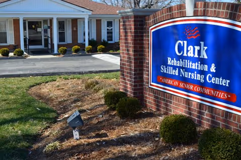 Exterior view of Clark Rehabilitation and Skilled Nursing Center showing the building entrance with white columns and a blue sign on a brick base displaying the facility name and American Senior Communities branding.