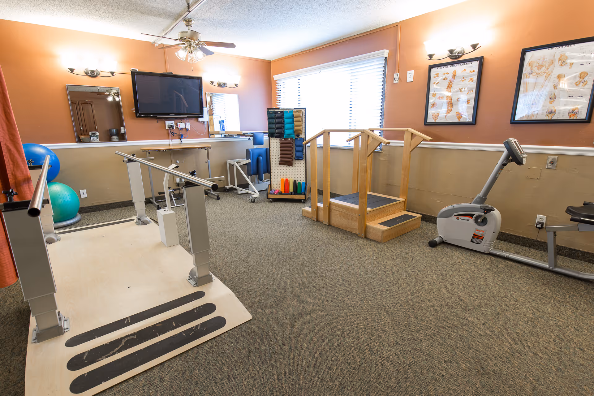 A senior therapy/exercise room with parallel bars, a small wooden step platform, an exercise bike and various fitness equipment.