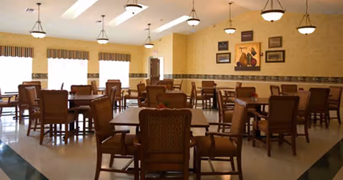 Spacious dining room with multiple wooden tables and chairs, warm yellow walls, pendant lighting, and framed artwork.