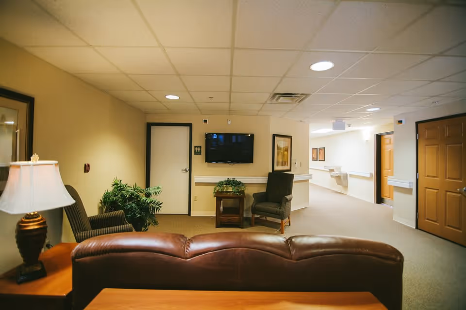 Interior view of a senior living facility lounge area with a brown leather couch in the foreground, two armchairs, a side table with a lamp, a potted plant, a wall-mounted TV, and a hallway with doors and handrails.