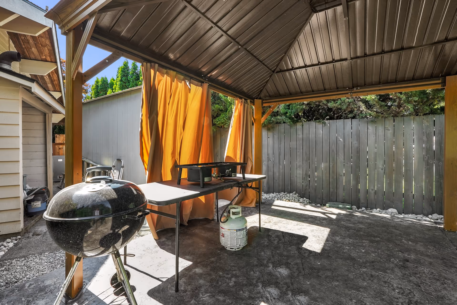 Covered outdoor patio area with a black charcoal grill on the left and a folding table holding a propane burner with a propane tank underneath. The patio has a metal roof and orange curtains hanging on one side. There is a wooden fence and some greenery in the background.