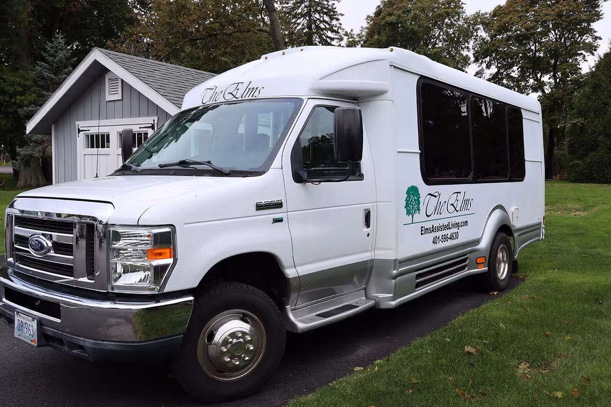 A white shuttle van labeled "The Elms" parked on a driveway beside a small gray outbuilding and lawn.