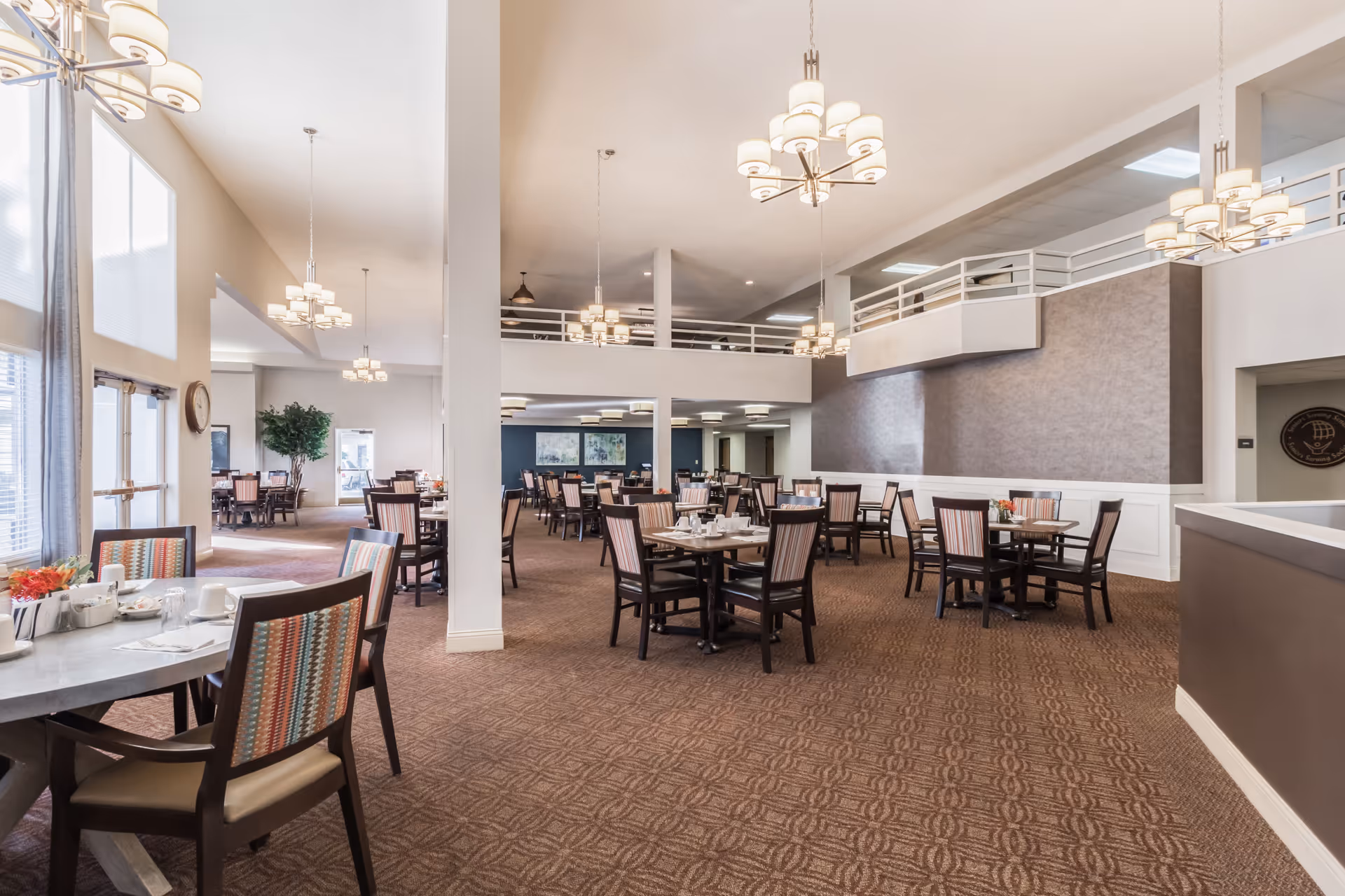 Spacious dining room in a senior living facility with multiple tables and chairs arranged neatly. The room features high ceilings, modern chandeliers, large windows allowing natural light, and a carpeted floor. The walls are painted in neutral tones with some decorative elements.