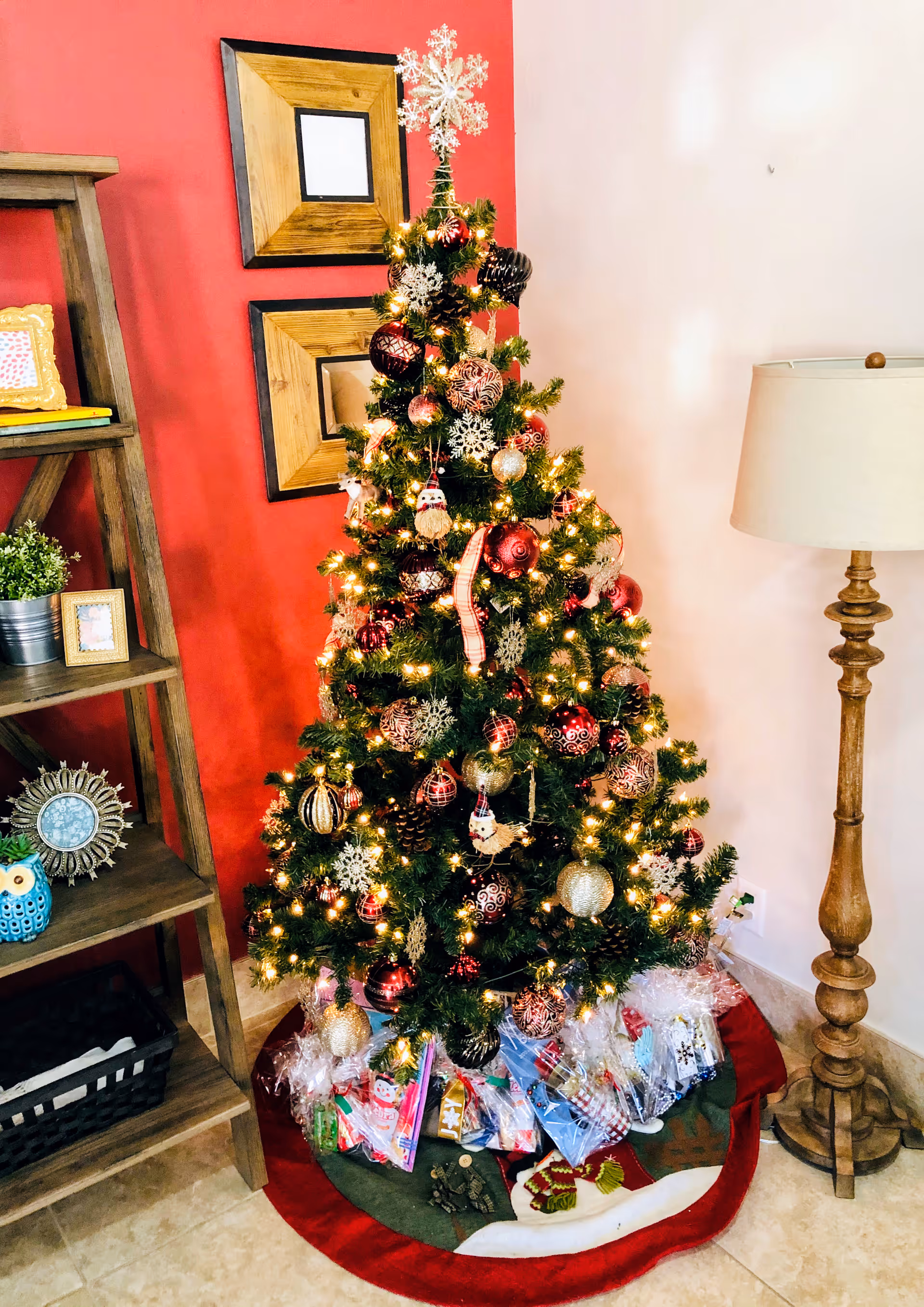 A decorated Christmas tree with lights, red and gold ornaments, and snowflake decorations stands in a corner of a room. Wrapped gifts are placed under the tree on a festive tree skirt. To the left of the tree is a wooden shelf with framed photos, a small plant, and decorative items. To the right is a wooden floor lamp with a beige lampshade. The wall behind the tree is painted red on one side and light pink on the other, with two wooden framed pictures hanging on the red wall.