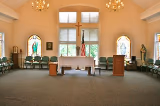 Interior of a chapel or worship room with a central altar covered with a white cloth, a wooden cross on the wall behind it, an American flag, stained glass windows on either side, green chairs arranged along the walls, and chandeliers hanging from the ceiling.