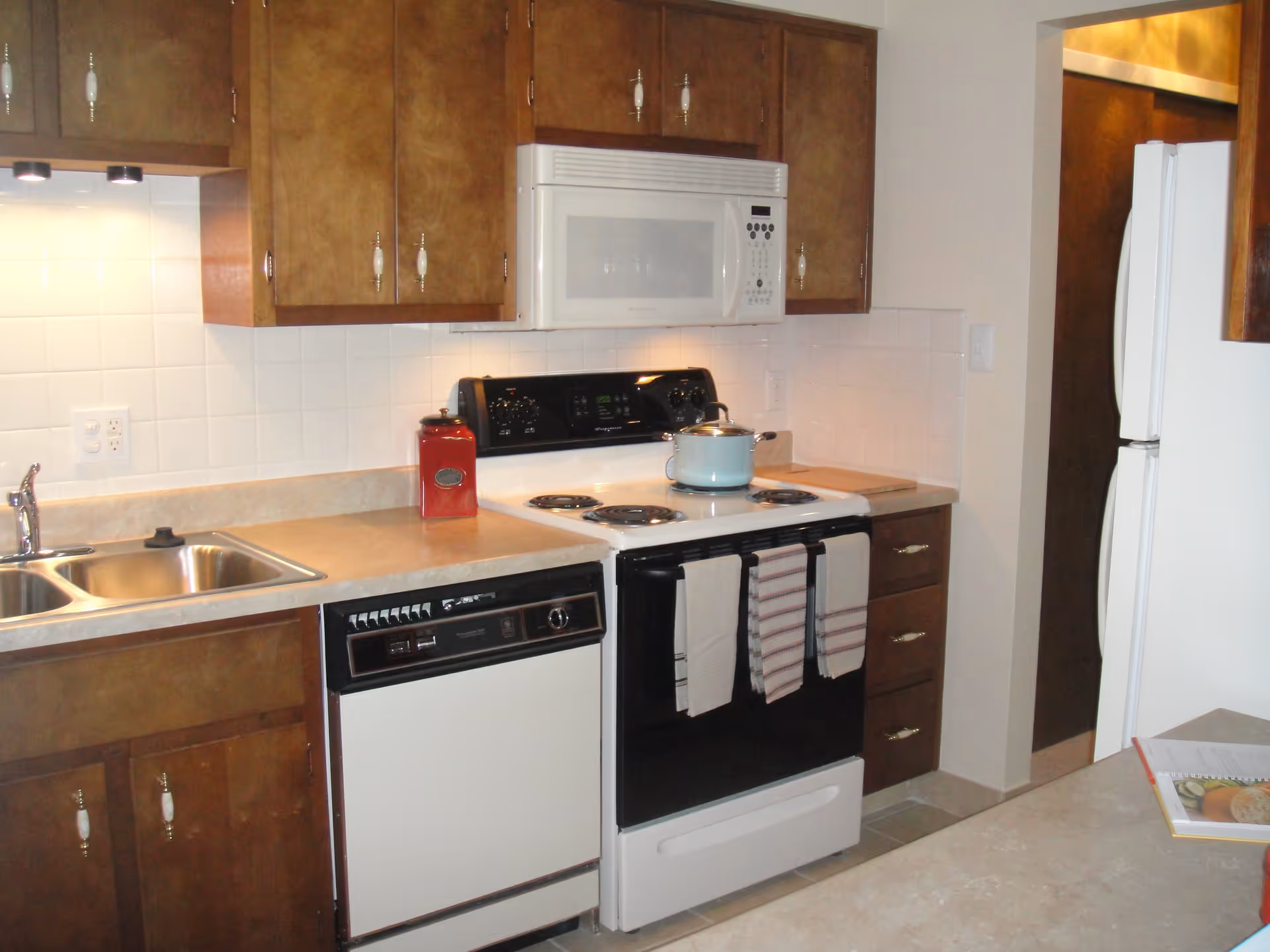 A kitchen with wooden cabinets, a white electric stove with a pot on it, a white microwave above the stove, a dishwasher, a double sink, and a white refrigerator. There are three dish towels hanging on the stove handle and a red container on the countertop.