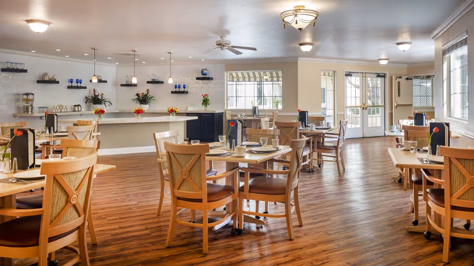 A spacious dining room with wooden floors and multiple tables set with plates, glasses, and menus. The room features wooden chairs with woven backs, ceiling fans, pendant lights, and large windows allowing natural light. There are decorative plants and shelves with glassware on the far wall.