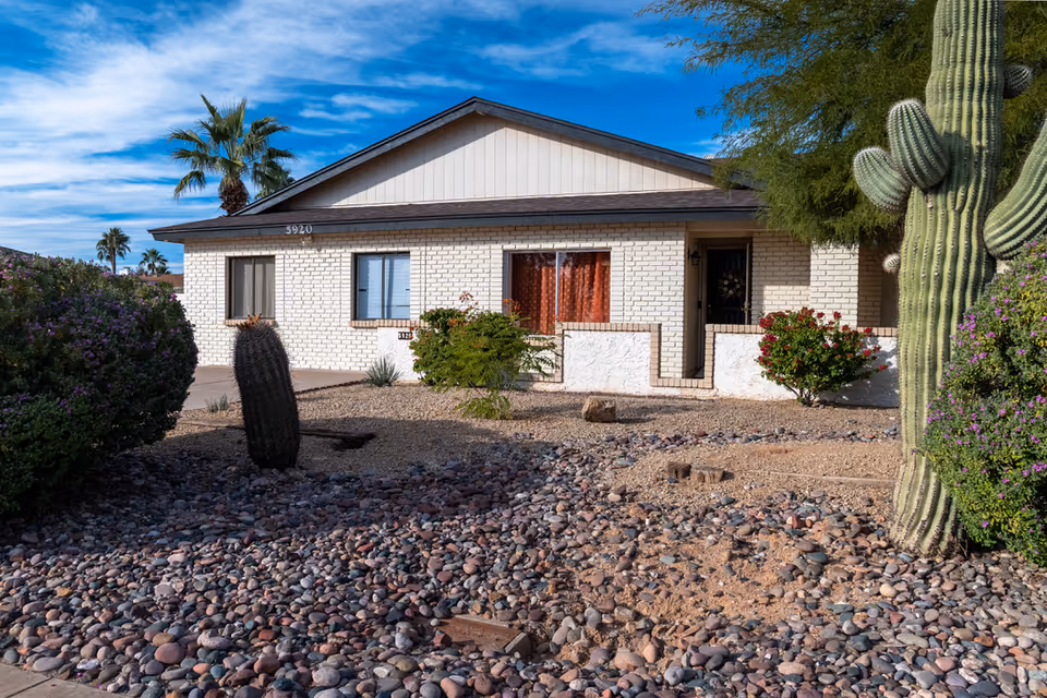 Front exterior view of a single-story white brick building with a brown roof, surrounded by desert landscaping including rocks, cacti, and bushes under a blue sky with some clouds.