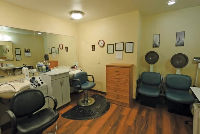 Interior of a salon or beauty care room with salon chairs, hair washing sinks, hair dryers, a wooden chest of drawers, and framed pictures and certificates on the walls.