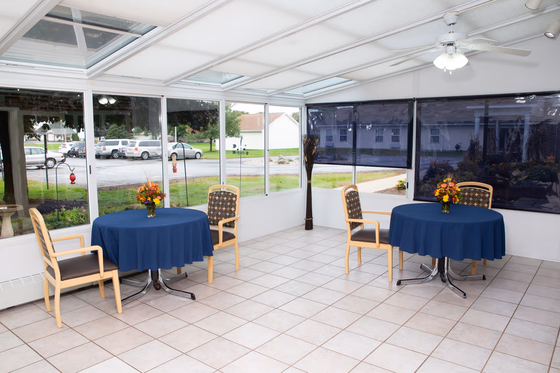 A bright sunroom with large windows and a glass ceiling, featuring two round tables covered with blue tablecloths. Each table has two wooden chairs with patterned cushions and a small vase of flowers. The floor is tiled, and there is a ceiling fan with lights above. Outside the windows, a parking lot and some greenery are visible.