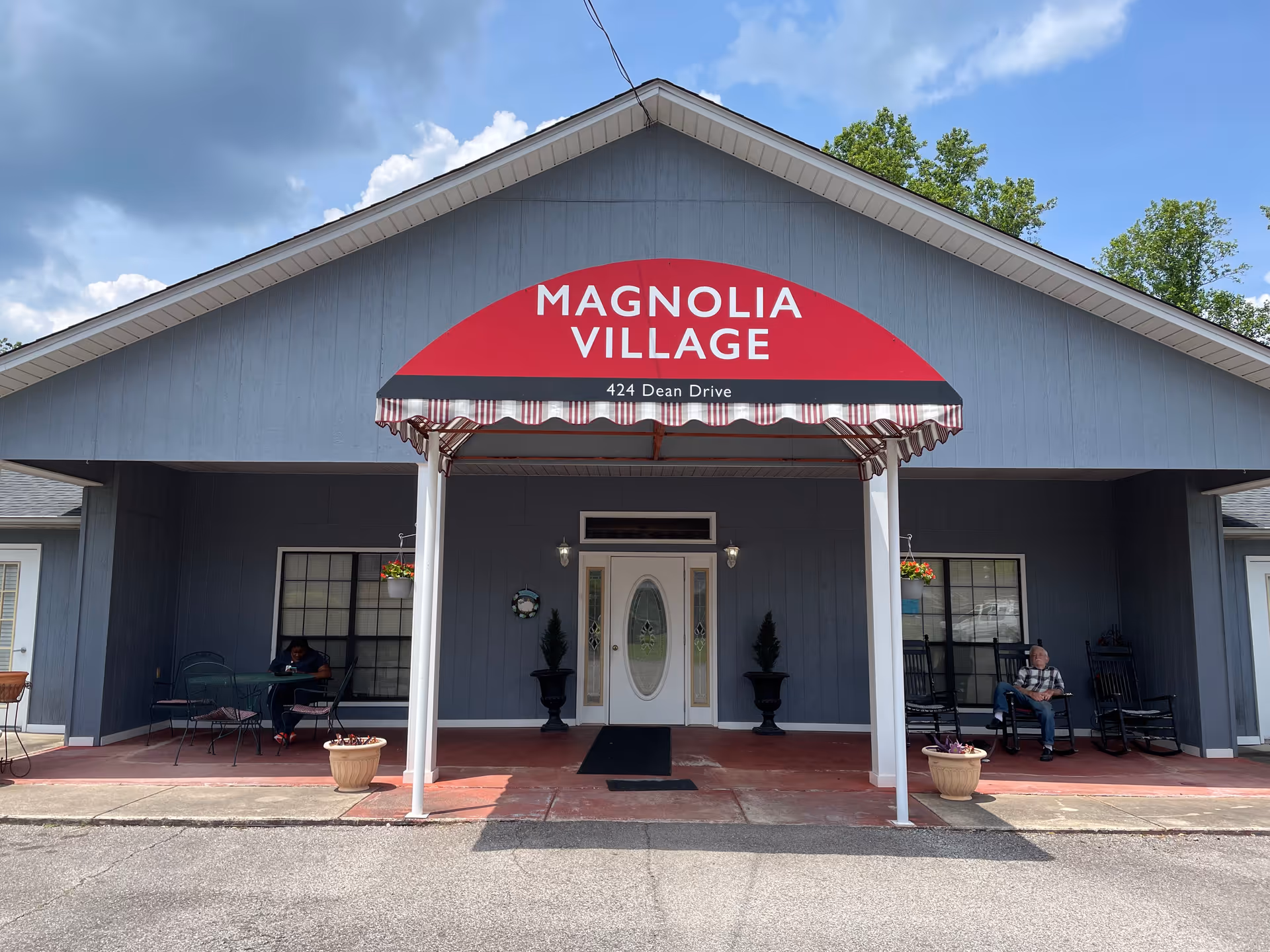 Front exterior view of Magnolia Village facility with a red awning displaying the name and address 424 Dean Drive. The building is gray with white trim, and there are two people sitting on either side of the entrance under covered porch areas with chairs and tables.