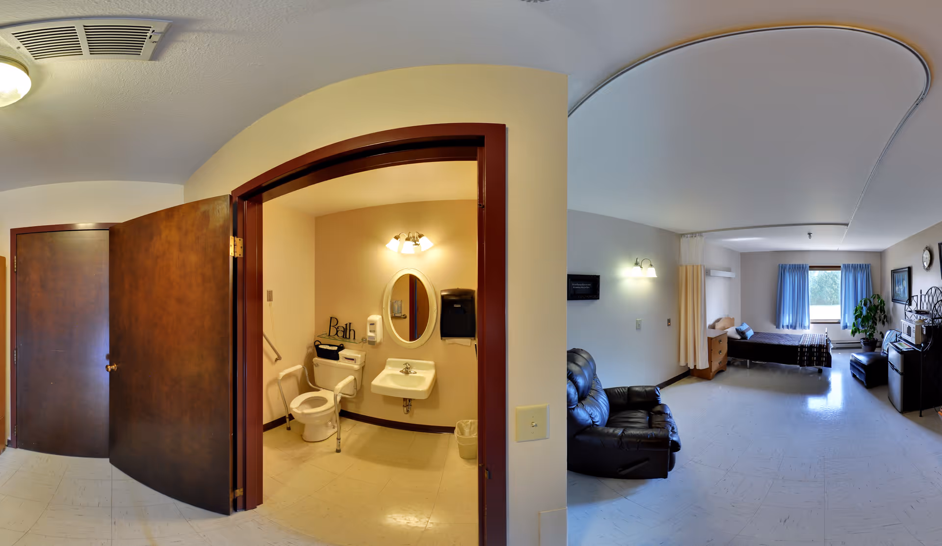 A senior living facility room with a visible bathroom and bedroom area. The bathroom has a toilet, sink with an oval mirror, soap dispenser, and grab bars. The bedroom area includes a bed, a leather recliner chair, a dresser, a small table, a window with blue curtains, and a potted plant near the window. The walls are light-colored, and the floor is tiled.
