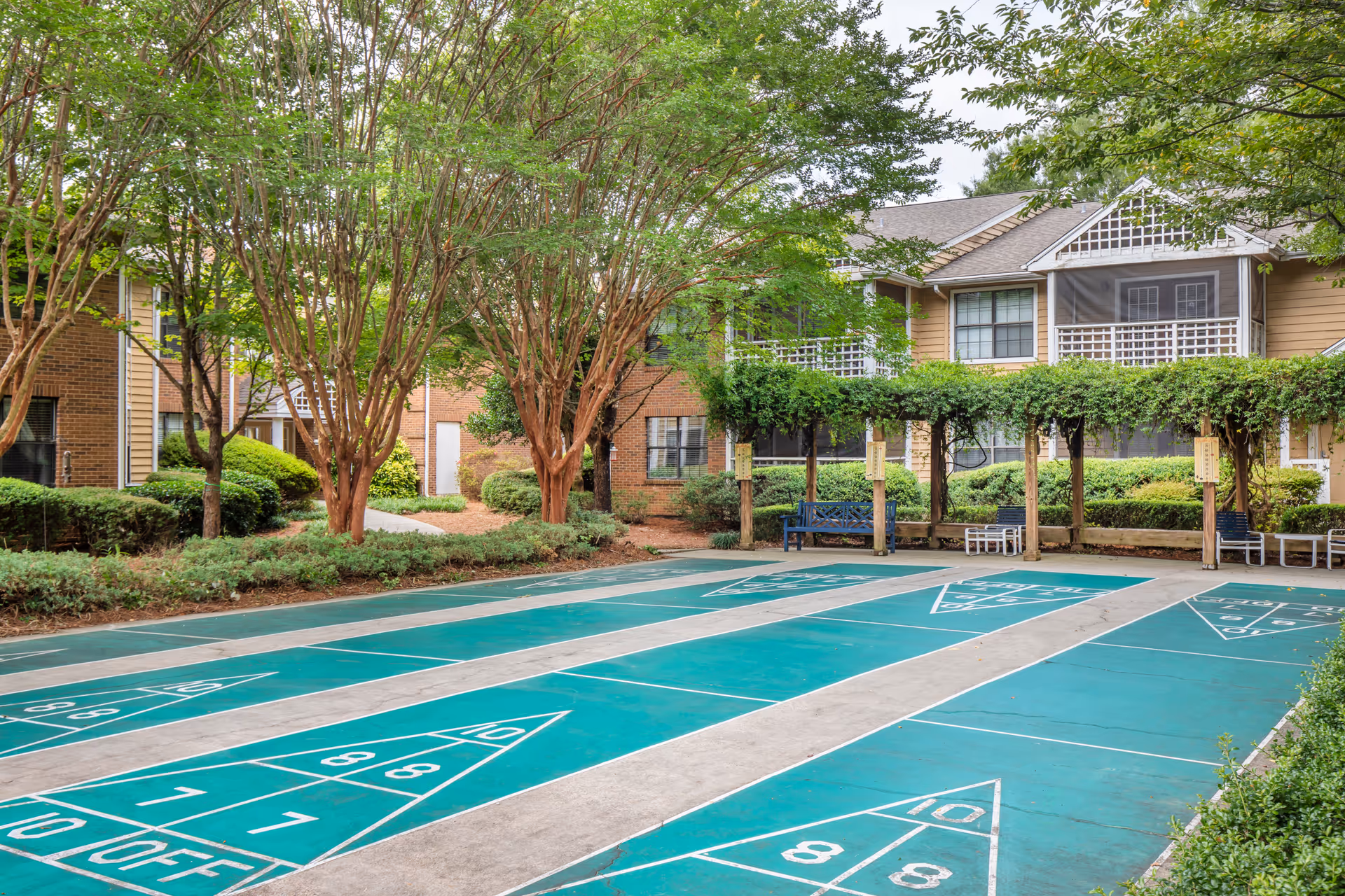 Shuffleboard courts in a landscaped courtyard with benches and a two-story residential building in the background.