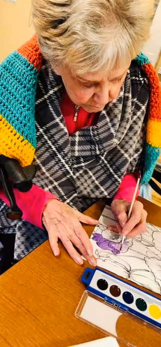 An elderly woman with short gray hair is sitting at a wooden table, painting a floral design on paper using watercolors. She is wearing a red shirt, a black and white plaid shawl, and a colorful crocheted blanket draped over her shoulders.