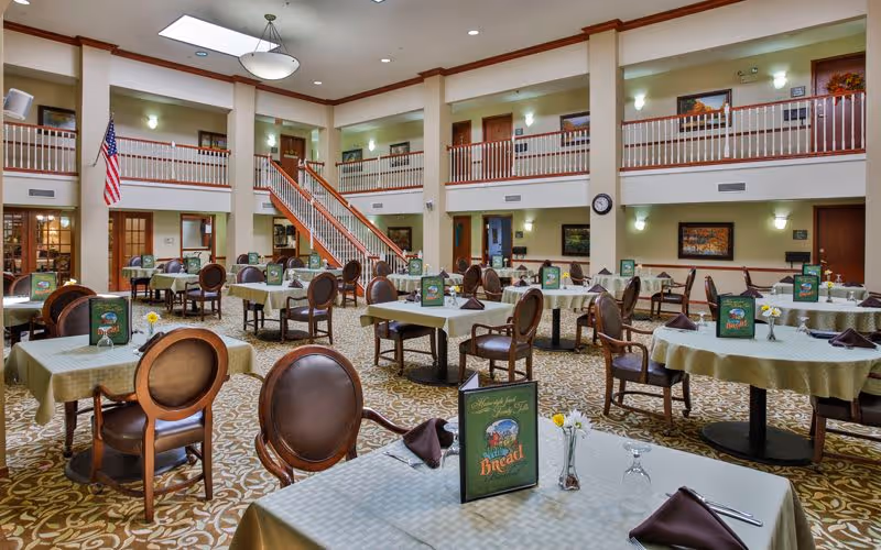 A spacious dining room in a senior living facility with multiple tables covered in light green tablecloths, each set with menus, napkins, and glassware. The room features a patterned carpet, wooden chairs with cushioned seats, a staircase leading to an upper level with railings, and an American flag displayed near the stairs. The walls are decorated with framed artwork and soft lighting fixtures.