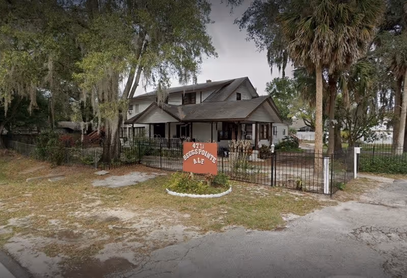 Exterior view of Beggs Pointe Assisted Living Facility, a two-story house-style building surrounded by trees and a black metal fence. There is a red sign in front with white text displaying the facility's name and address. The ground is a mix of grass and pavement.