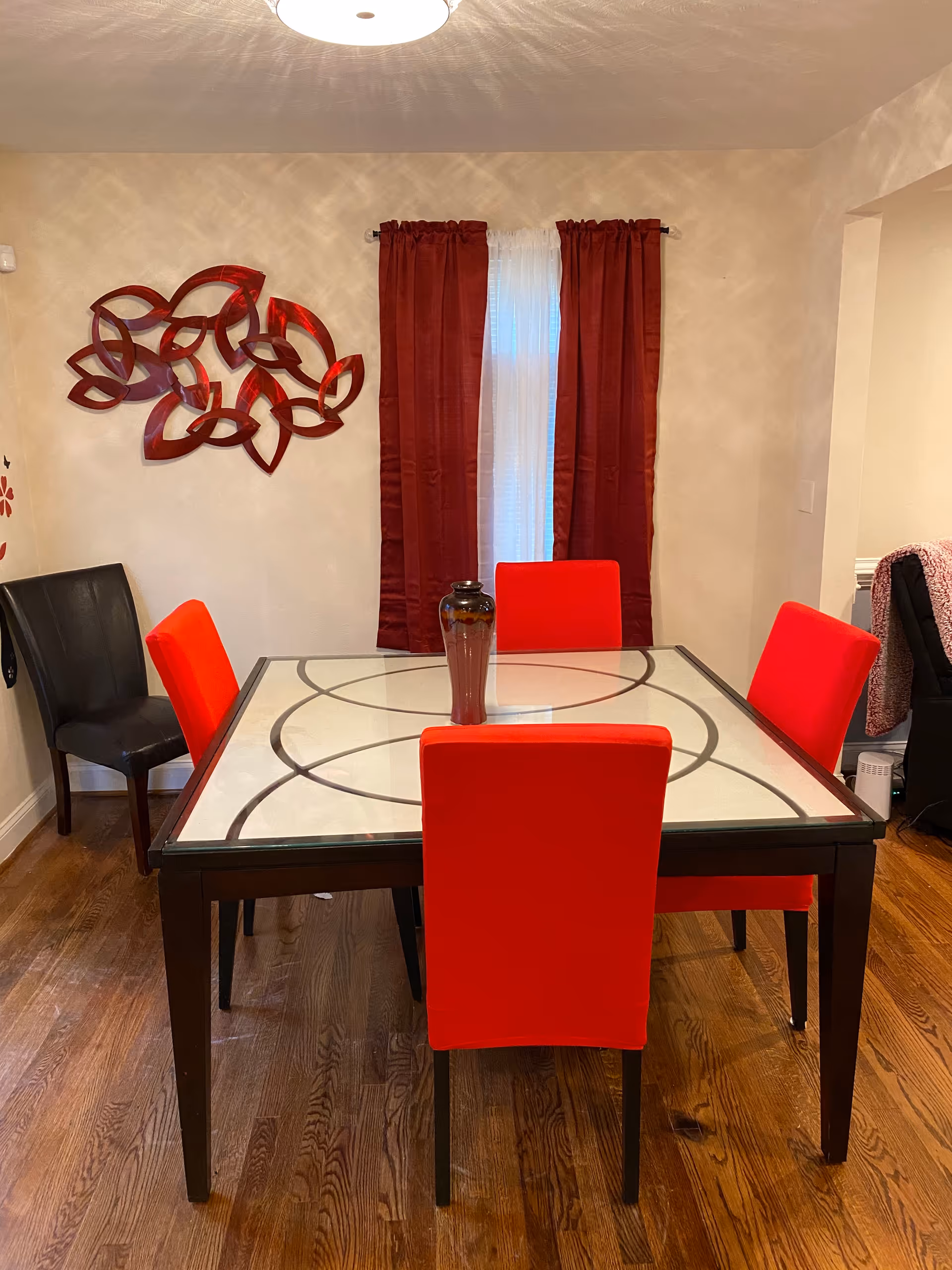 Dining room with a glass-top table surrounded by red chairs, a black chair, a central vase, red curtains and decorative wall art.
