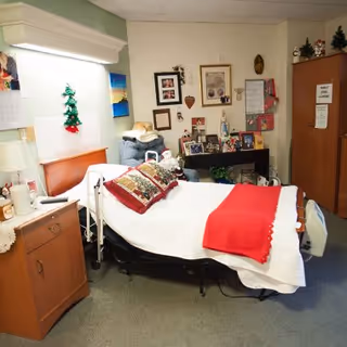 A cozy nursing facility room with a hospital bed covered in white and red bedding, including decorative pillows. The room has a wooden cabinet with a lamp and various personal items. The walls are decorated with framed pictures, a calendar, and holiday decorations. There is a comfortable chair next to the bed and a wooden wardrobe in the corner.