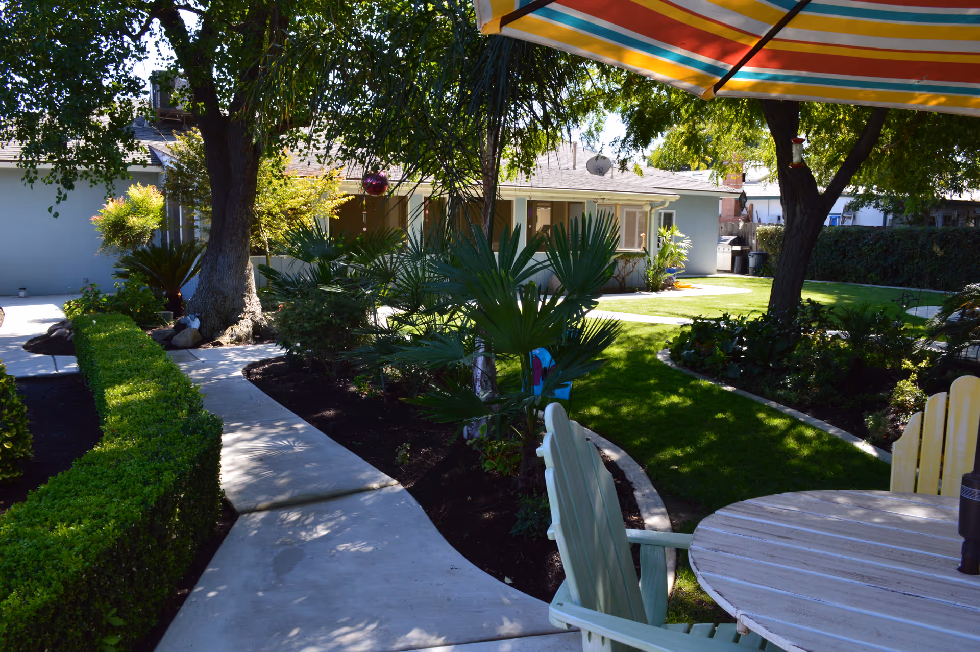 A shaded outdoor garden area with a concrete pathway winding through green bushes and trees. There is a round wooden table with chairs under a colorful striped umbrella in the foreground. A single-story building with windows and a satellite dish is visible in the background.