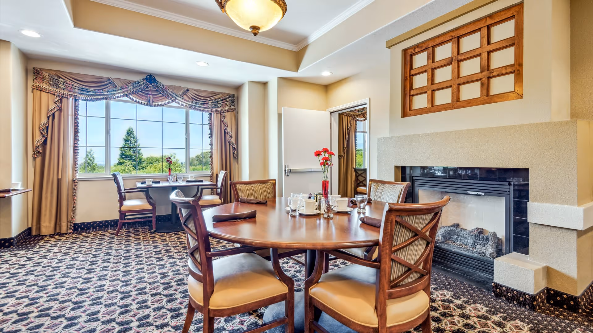 A cozy dining area with a round wooden table set with cups, glasses, and a vase with red flowers. Four cushioned chairs surround the table. The room features a large window with elegant drapes, patterned carpet, a fireplace, and a ceiling light fixture.