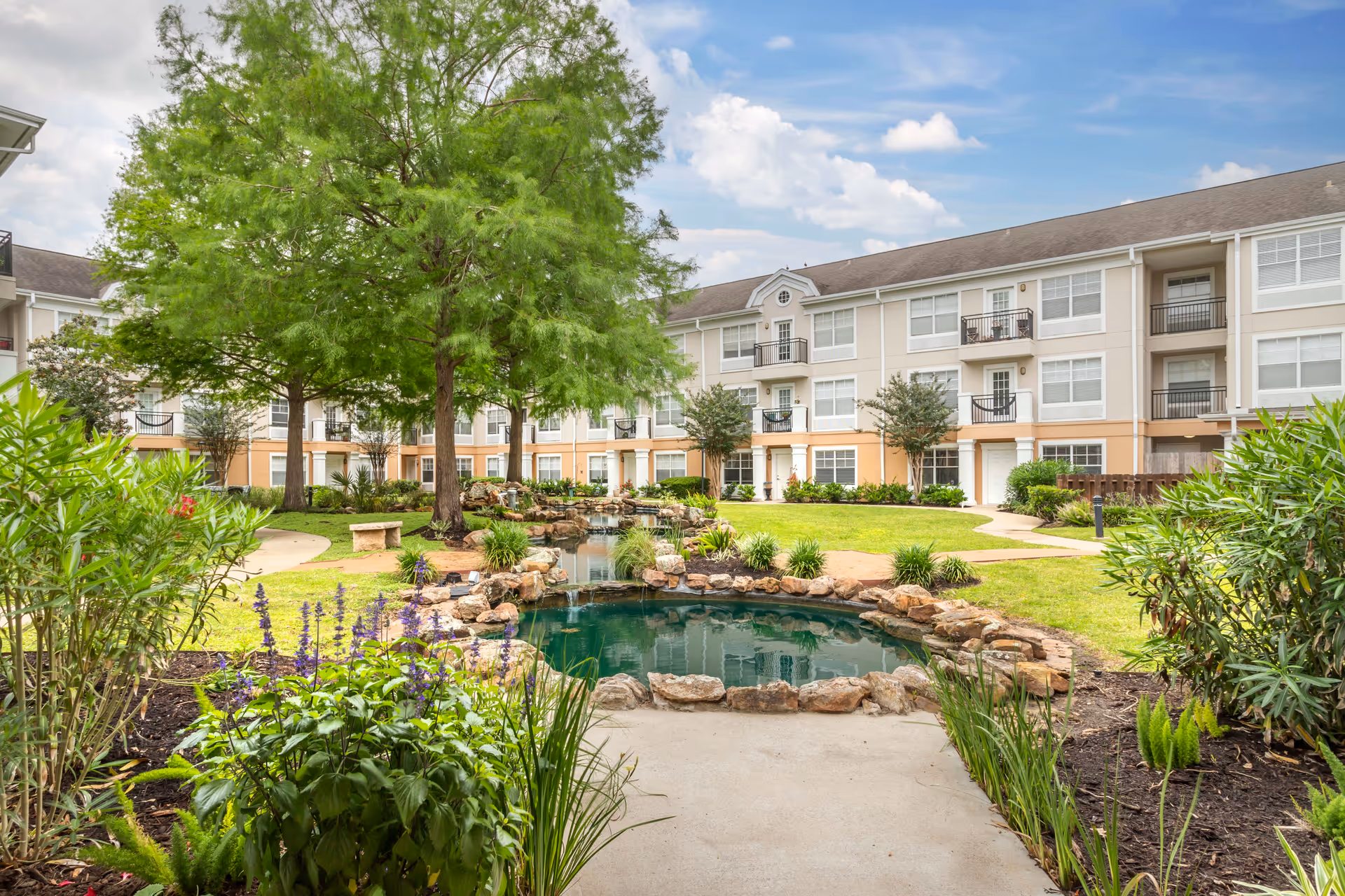 A landscaped outdoor courtyard area at Brookdale Clear Lake featuring a small pond with rocks around it, green grass, trees, shrubs, and a three-story residential building in the background under a partly cloudy sky.