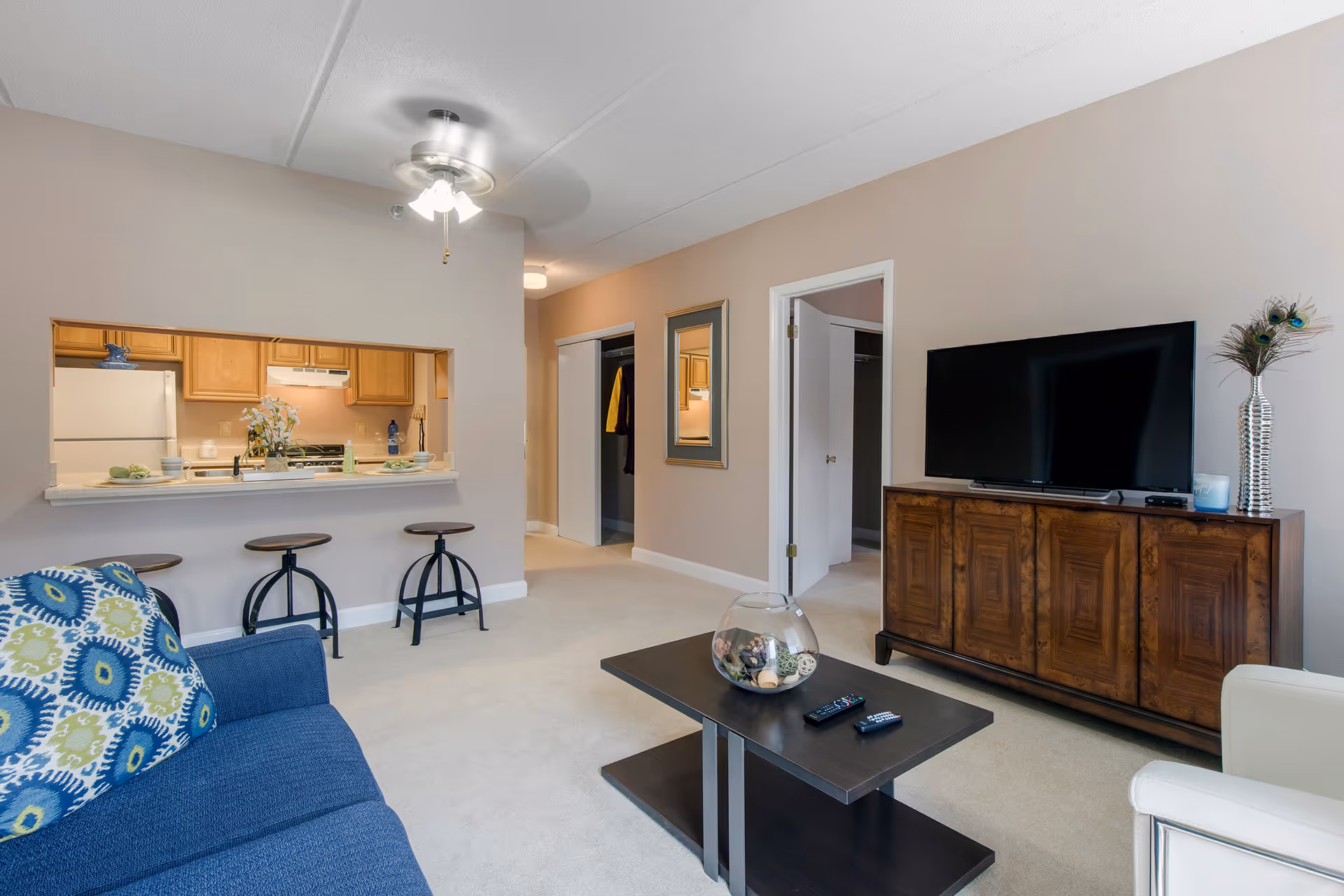 A cozy living room with a blue couch featuring patterned pillows, a dark wooden coffee table with decorative items and remote controls, a wooden TV stand with a flat-screen TV, and a silver vase with peacock feathers. The room has beige walls and carpet, a ceiling fan with lights, and an open pass-through window to a kitchen with wooden cabinets and white appliances. There are two open doorways and a closet with sliding doors visible in the background.